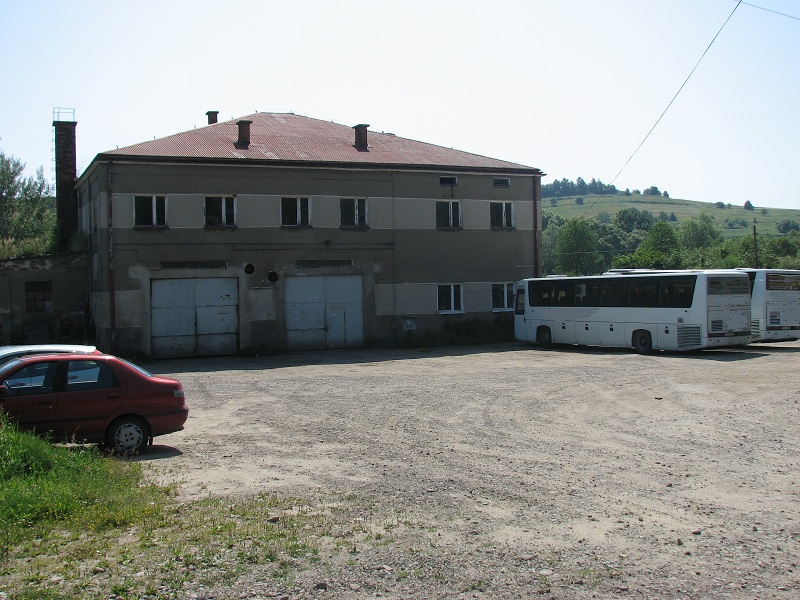 Dwu kondygnacyjny budynek z garażami, ceglanym kominem i czerwonym dachem. Przed budynkiem zaparkowane autobusy i samochód osobowy.